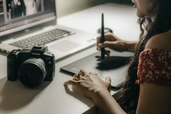 A person uses a stylus to work on a graphic tablet beside a laptop. A digital camera is also placed on the table, indicating a creative workspace. The focus is on the hand holding the stylus, illustrating an active engagement in a digital design or photo editing task.