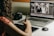 A person is working at a desk using a laptop and a graphics tablet with a stylus. A digital photo editing software is open on the laptop, displaying a black and white image of a wedding couple. A professional camera rests on the desk next to the laptop.
