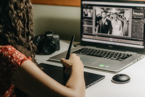 A person is working at a desk using a laptop and a graphics tablet with a stylus. A digital photo editing software is open on the laptop, displaying a black and white image of a wedding couple. A professional camera rests on the desk next to the laptop.