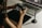 A person is holding a digital camera and examining its settings while sitting at a desk. In front of them, there is a laptop displaying photo editing software, a graphics tablet, and a stylus pen. The lighting is soft and warm, creating a focused and creative atmosphere.
