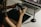 A person is holding a digital camera and examining its settings while sitting at a desk. In front of them, there is a laptop displaying photo editing software, a graphics tablet, and a stylus pen. The lighting is soft and warm, creating a focused and creative atmosphere.