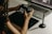 A person is holding a digital camera and examining its settings while sitting at a desk. In front of them, there is a laptop displaying photo editing software, a graphics tablet, and a stylus pen. The lighting is soft and warm, creating a focused and creative atmosphere.