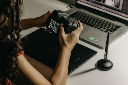 A person is holding a digital camera and examining its settings while sitting at a desk. In front of them, there is a laptop displaying photo editing software, a graphics tablet, and a stylus pen. The lighting is soft and warm, creating a focused and creative atmosphere.