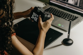 A person is holding a digital camera and examining its settings while sitting at a desk. In front of them, there is a laptop displaying photo editing software, a graphics tablet, and a stylus pen. The lighting is soft and warm, creating a focused and creative atmosphere.