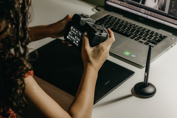 A person is holding a digital camera and examining its settings while sitting at a desk. In front of them, there is a laptop displaying photo editing software, a graphics tablet, and a stylus pen. The lighting is soft and warm, creating a focused and creative atmosphere.
