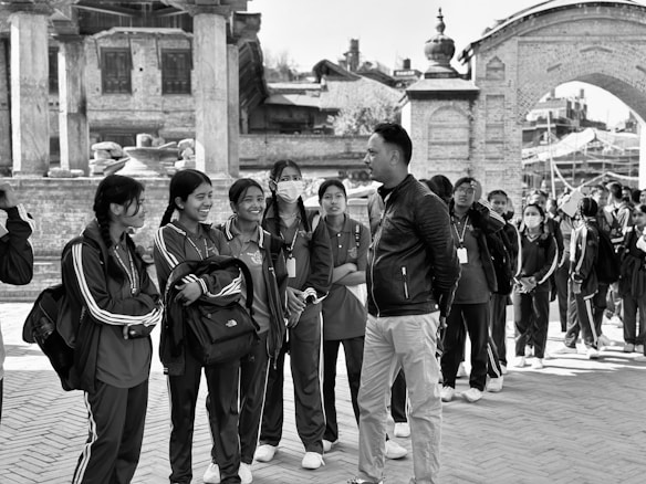 A group of young people wearing matching athletic clothes are gathered in a line, talking and smiling amongst each other. A man stands facing them, engaging in conversation. The backdrop features historic architecture, including stone columns and brick structures.