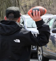A person wearing a black jacket featuring a North Face logo and a black cap is holding a black and orange football. They are standing outdoors near parked cars and trees, possibly in a park or parking lot setting.