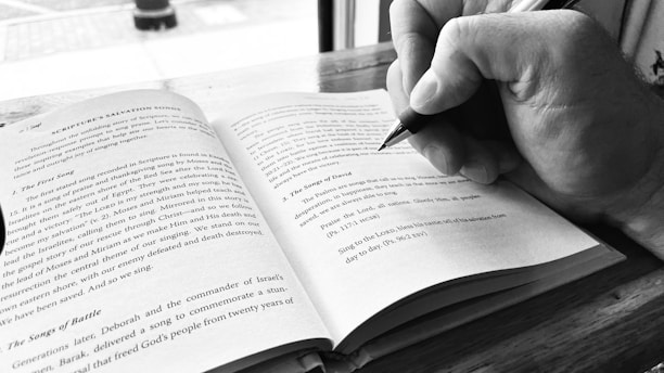 Close-up of a hand writing notes next to a vintage leather-bound book and a pair of glasses, sunlight streaming in.