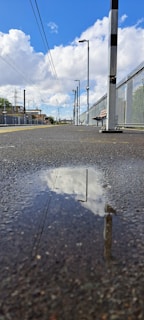 A railway platform with a long, empty stretch of pavement leading to the horizon. Overhead power lines run parallel to the platform, and there is a large puddle on the ground reflecting the blue sky and white clouds. Metal fencing lines one side of the platform, accompanied by lampposts and a solitary bench.