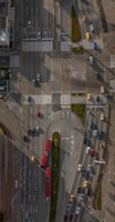 An aerial view of a busy urban intersection with multiple lanes of traffic, featuring various vehicles such as cars, buses, and taxis. The roads are marked with lane indicators and pedestrian crossings. There are sections of greenery, and the area is well-structured with clear signage. Shadows cast from nearby structures create patterns on the road.