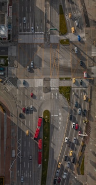 An aerial view of a busy urban intersection with multiple lanes of traffic, featuring various vehicles such as cars, buses, and taxis. The roads are marked with lane indicators and pedestrian crossings. There are sections of greenery, and the area is well-structured with clear signage. Shadows cast from nearby structures create patterns on the road.