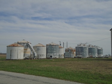 A collection of weathered, metal grain silos with rusty tops is situated on a field. The sky above is partly cloudy, creating a peaceful rural landscape. The grass surrounding the silos is a mixture of green and brown, indicating a transition period between seasons.