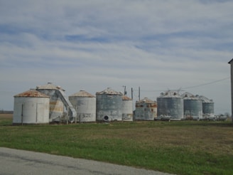 A collection of weathered, metal grain silos with rusty tops is situated on a field. The sky above is partly cloudy, creating a peaceful rural landscape. The grass surrounding the silos is a mixture of green and brown, indicating a transition period between seasons.