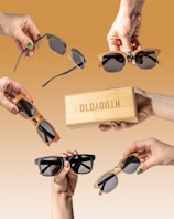 Multiple hands holding various styles of sunglasses and a wooden box against a brown gradient background. Each hand presents a unique design, showcasing a range of frame colors and shapes, highlighting the sunglasses collection.