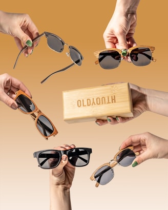 Multiple hands holding various styles of sunglasses and a wooden box against a brown gradient background. Each hand presents a unique design, showcasing a range of frame colors and shapes, highlighting the sunglasses collection.
