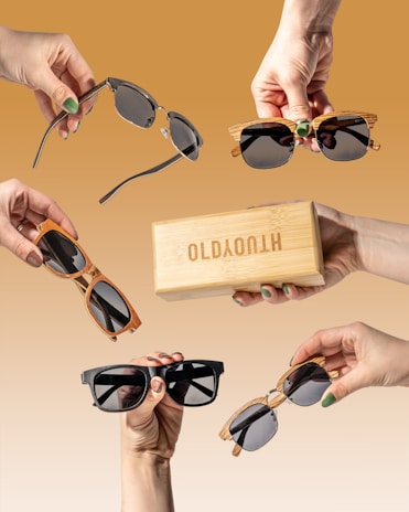 Multiple hands holding various styles of sunglasses and a wooden box against a brown gradient background. Each hand presents a unique design, showcasing a range of frame colors and shapes, highlighting the sunglasses collection.