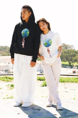 Two people stand outdoors on a sunny day, dressed in casual attire. One person wears a black hoodie with a graphic of a basketball, and white pants. The other person wears a white T-shirt with the same graphic and light-colored pants. They are standing close to each other, with one person leaning slightly on the other. The background includes greenery and distant buildings.