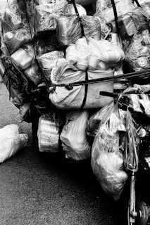 A heavily loaded bicycle carrying numerous plastic bags filled with various items, all secured with black straps. The collection of bags appears to contain recyclables or items for transport, arranged in a way that almost obscures the bicycle itself. The image is in black and white, emphasizing the textures and shapes of the plastic and fabric.