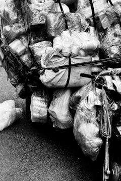 A heavily loaded bicycle carrying numerous plastic bags filled with various items, all secured with black straps. The collection of bags appears to contain recyclables or items for transport, arranged in a way that almost obscures the bicycle itself. The image is in black and white, emphasizing the textures and shapes of the plastic and fabric.