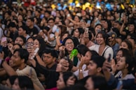 A large crowd of people gather closely together, many of them holding up smartphones to capture moments. The group appears to be at an event, possibly a concert, as they are focused on something beyond the frame. The expressions are joyful and enthusiastic.