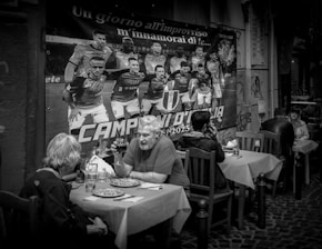 People dining at outdoor tables in front of a large poster showing a soccer team with the text 'Campioni d'Italia' and the year 2023. The scene appears to be in an urban alley or street, with a cobblestone pavement and graffiti on the walls.