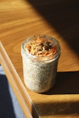 A glass jar filled with chia pudding is placed on a wooden surface. The pudding is topped with shredded carrots and granola, creating a layered, textured appearance. The sunlight casts a warm glow, highlighting the ingredients against the wooden table.