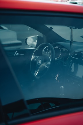 The interior of a car is visible through the window, highlighting a Volkswagen steering wheel, the dashboard, and part of the front seat. The vehicle's interior appears to be modern and well-maintained, and the glass reflects some exterior light.