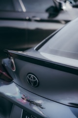A close-up view of the rear end of a gray Toyota Supra car, focusing on the emblem and the logo. Another car is partially visible in the background, slightly out of focus.