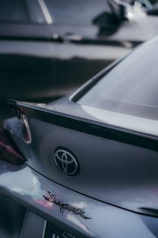A close-up view of the rear end of a gray Toyota Supra car, focusing on the emblem and the logo. Another car is partially visible in the background, slightly out of focus.
