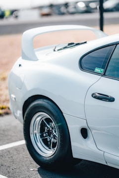 A close-up of the rear side of a white sports car featuring a prominent spoiler and sleek design. The car is parked on a paved lot with blurred vehicles in the background. The tire is glossy and the window has a sticker with the word 'REAL' visible.