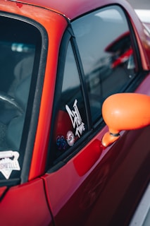 A close-up view of a red car, focusing on the side window and the rear-view mirror. The window displays several stickers, one of which reads 'Get Set Time'. The car's body and side mirror show a strong red and orange palette.