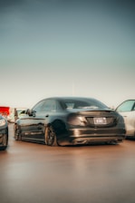 A sleek black luxury sedan with tinted windows is parked on a smooth surface. The rear view of the car is prominently displayed, showcasing its elegant design and shiny exterior. Two other cars are partially visible on either side, and there is a clear sky above. Some people are seen in the distant background.