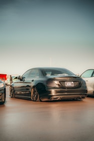 A sleek black luxury sedan with tinted windows is parked on a smooth surface. The rear view of the car is prominently displayed, showcasing its elegant design and shiny exterior. Two other cars are partially visible on either side, and there is a clear sky above. Some people are seen in the distant background.