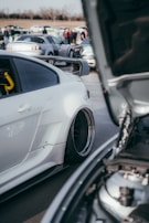 A car meet with several modified cars parked close together. The focus is on a white sports car with a large rear wing and custom rims. The hood of another car in the foreground is open, revealing some of its engine components. People in the background are gathered and conversing, with more cars parked behind.