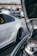 A car meet with several modified cars parked close together. The focus is on a white sports car with a large rear wing and custom rims. The hood of another car in the foreground is open, revealing some of its engine components. People in the background are gathered and conversing, with more cars parked behind.
