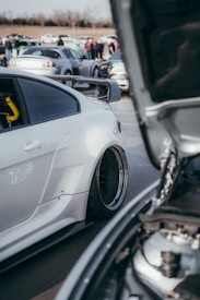 A car meet with several modified cars parked close together. The focus is on a white sports car with a large rear wing and custom rims. The hood of another car in the foreground is open, revealing some of its engine components. People in the background are gathered and conversing, with more cars parked behind.