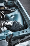 A close-up view of an automotive engine bay featuring a high-performance air intake system with a chrome and black finish. The car's body is painted light blue, and there are various metal components visible, including tubing and wiring.