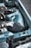 A close-up view of an automotive engine bay featuring a high-performance air intake system with a chrome and black finish. The car's body is painted light blue, and there are various metal components visible, including tubing and wiring.