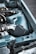A close-up view of an automotive engine bay featuring a high-performance air intake system with a chrome and black finish. The car's body is painted light blue, and there are various metal components visible, including tubing and wiring.