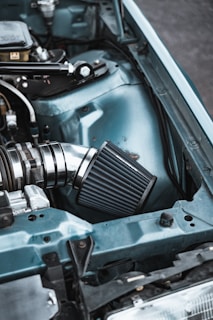 A close-up view of an automotive engine bay featuring a high-performance air intake system with a chrome and black finish. The car's body is painted light blue, and there are various metal components visible, including tubing and wiring.