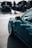A close-up view of a sleek, modern car with polished white rims and a teal body. In the background, a group of people is gathered around other cars with their hoods open, likely at a car show or meetup. The scene suggests a focus on automotive enthusiasm and technical inspection.
