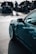A close-up view of a sleek, modern car with polished white rims and a teal body. In the background, a group of people is gathered around other cars with their hoods open, likely at a car show or meetup. The scene suggests a focus on automotive enthusiasm and technical inspection.