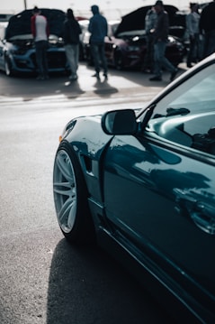 A close-up view of a sleek, modern car with polished white rims and a teal body. In the background, a group of people is gathered around other cars with their hoods open, likely at a car show or meetup. The scene suggests a focus on automotive enthusiasm and technical inspection.