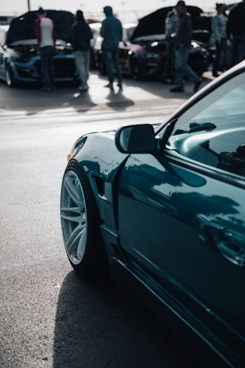 A close-up view of a sleek, modern car with polished white rims and a teal body. In the background, a group of people is gathered around other cars with their hoods open, likely at a car show or meetup. The scene suggests a focus on automotive enthusiasm and technical inspection.