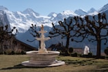 A beautiful landscape featuring a snow-capped mountain range in the background, with a well-maintained garden in the foreground. The garden has a large decorative fountain as its centerpiece surrounded by neatly trimmed bushes and pruned trees with bare branches. A few traditional wooden houses are visible in the middle ground, adding a quaint charm to the scene.