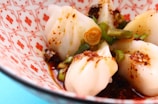 A close-up of Asian-style dumplings garnished with sliced green onions and chili flakes. They are served in a decorative bowl with red patterns on a white background.