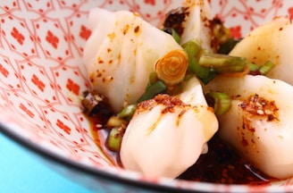 A close-up of Asian-style dumplings garnished with sliced green onions and chili flakes. They are served in a decorative bowl with red patterns on a white background.