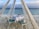 A romantic beachfront dining setup under a wooden canopy with sheer curtains. A table with a white tablecloth is neatly set with two chairs featuring turquoise cushions, a decorative centerpiece, and a folded napkin. The calm ocean is visible in the background, under a partly cloudy sky.