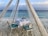 A romantic beachfront dining setup under a wooden canopy with sheer curtains. A table with a white tablecloth is neatly set with two chairs featuring turquoise cushions, a decorative centerpiece, and a folded napkin. The calm ocean is visible in the background, under a partly cloudy sky.