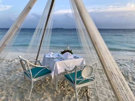 A romantic beachfront dining setup under a wooden canopy with sheer curtains. A table with a white tablecloth is neatly set with two chairs featuring turquoise cushions, a decorative centerpiece, and a folded napkin. The calm ocean is visible in the background, under a partly cloudy sky.